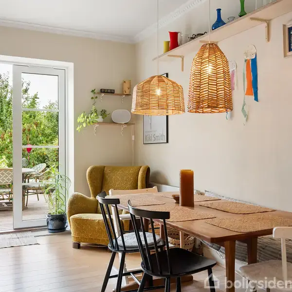 Et spisebord med stole står i et lyst rum, hvor to flettede lamper hænger over. Døre åbner ud til en terrasse med planteudsmykning og havemøbler.