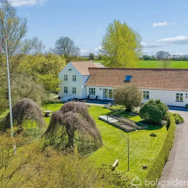 Et langt, hvidt hus med rødt tegltag står omkranset af frodige træer og en velplejet have. En grusvej leder op til en parkeret bil i gårdspladsen.