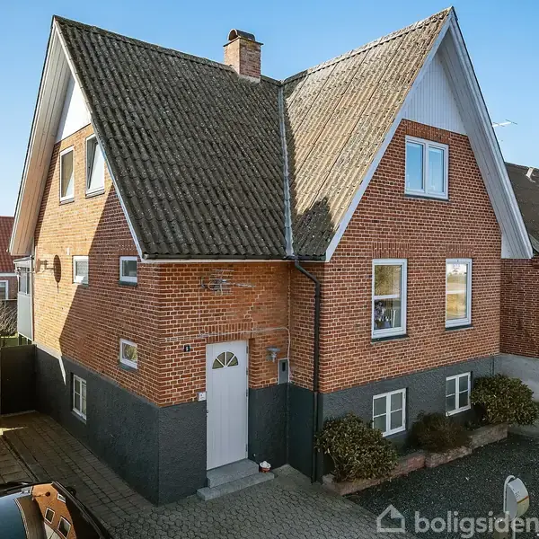 Et rødt murstenshus med højt gavltag står stille i et boligområde, omgivet af andre huse og en flisesti foran. En carport og en bil er til højre.