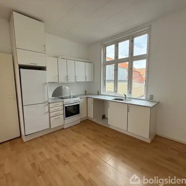Køkken med hvide skabe og apparater, herunder en ovn og køleskab, står i et rum med trægulv. Store vinduer lader lys ind fra en baggård med røde tage.