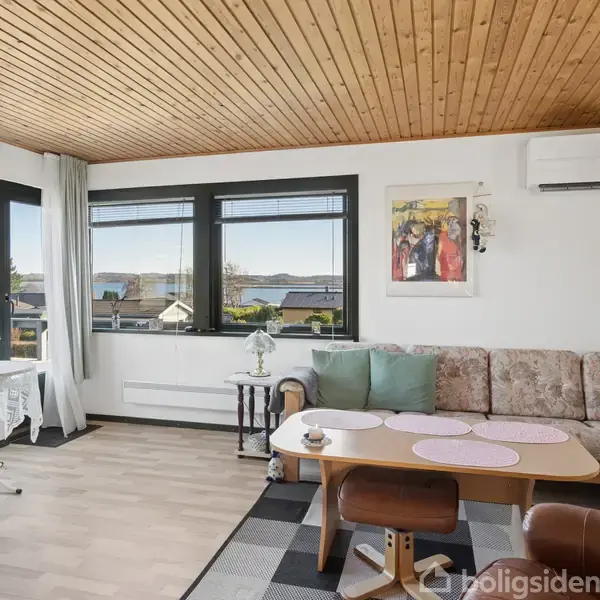 Stue med brun sofa mod hvid væg, lakeret træloft, og bord med stole. Store vinduer viser udsigt til huse og bjerge. Aircondition er monteret over sofaen.