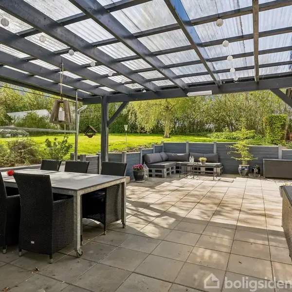 Terrasse med glasoverdækning, spisebord med stole, hjørnesofa og planter. Beliggende i en have med grønne buske og et træ i baggrunden.