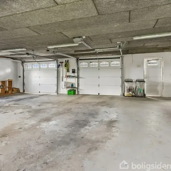 Garage med tre store porte, opbevaringshylder og cykler, der står op mod væggene. Gulvet er slidt, og der er loftslamper. Miljøet er rummeligt med hvidmalede vægge.