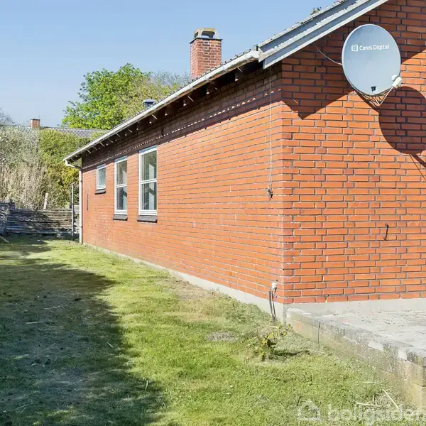 Et rødt murstenshus med en hvid dør og vinduer på siden. En parabol er monteret på væggen. Området har en græsplæne med få buske og træer i baggrunden.