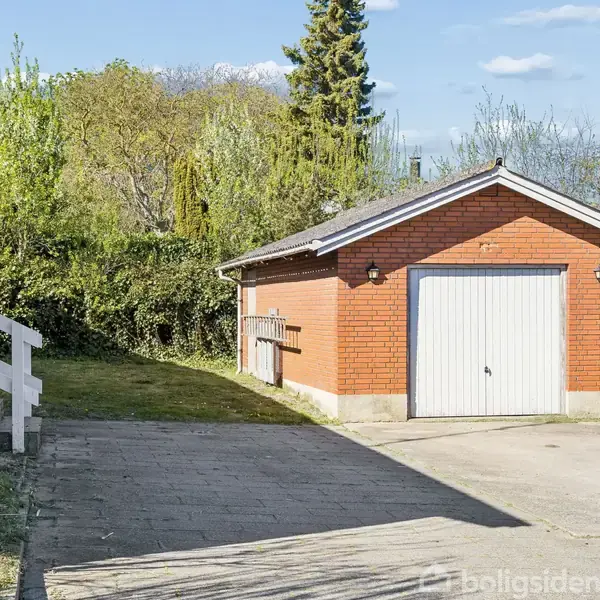 Garage i rød mursten med hvid port, står stille ved siden af en have med buske og træer. Beliggende ved et hus med trætrappe og brolagt indkørsel.