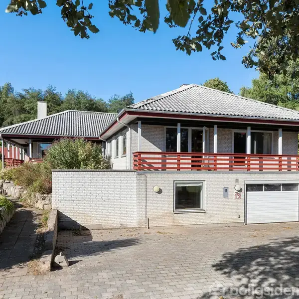 Hus med hvid murstensfacade og rødt træværk står på en brolagt indkørsel foran en garageport. Omgivet af grønne træer og buske i en stille boligkvarter.