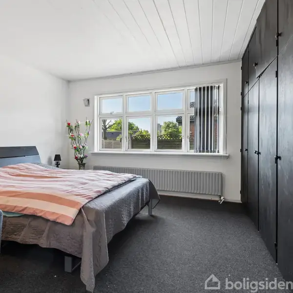 Soveværelse med en dobbeltseng dækket af en stribet dyne, placeret ved en stor vindueskarm med udsigt. Skabet står til højre; en grå sofa står nær sengen.