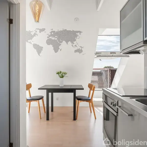 Køkken med moderne apparater; spisebord med to stole, vase med blomster; verdenskort på væggen; lysindfald fra stort vindue; hvid og lys træfarvet indretning.