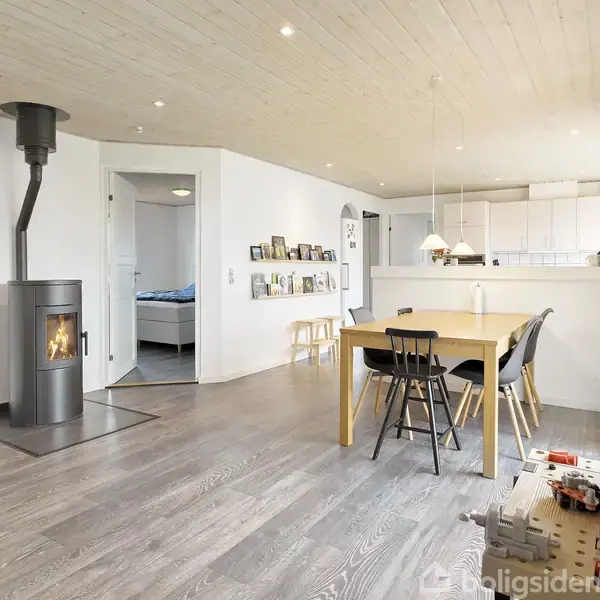 Brændeovn står i stuehjørnet og er tændt, mens et spisebord med stole er centralt placeret. Køkkenet ligger i baggrunden med hvide skabe og åbne hylder.