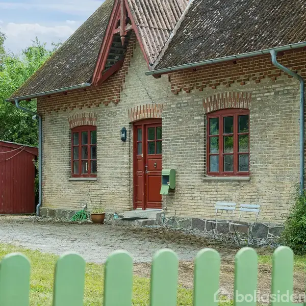 En murstensbygning med rødt tag og dør står på en gårdsplads bag et grønt hegn. Omkring huset ses buske og træer. Husnummeret 34 er synligt ved døren.