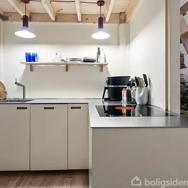 Et køkken med hvidt skab og sort bordplade. En vask og kogeplade ses. Hylde med glas og tallerkener hænger over bordpladen. Trætrappe er i baggrunden under loftsbelysning.