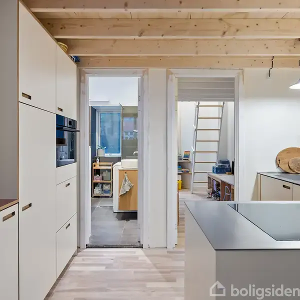 Køkken med lyse trægulve og træloft, indbygningsskabe til venstre, køkkenø i forgrunden, og åben dør mod stue med stige til højre. Lampe hænger fra loftet.