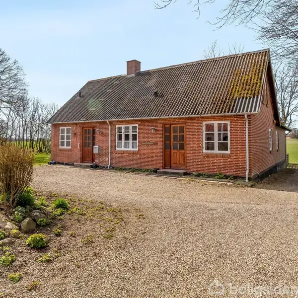 Et rødt murstenshus med to døre og et skrånende tag står på en grusvej. Omgivet af træer og åbne marker, under en blå himmel.