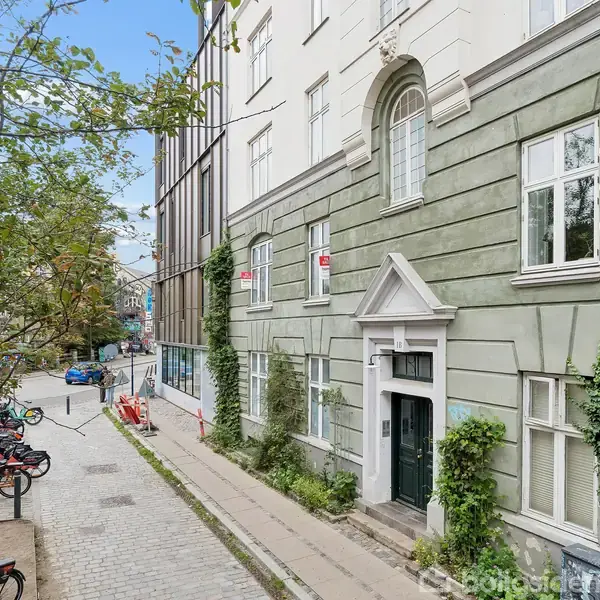 Grøn bygning med hvide vinduer står langs en brostensbelagt gade. Foran bygningen er cykler parkeret nær fortovet. Træer og buske vokser langs facaden og gaden.