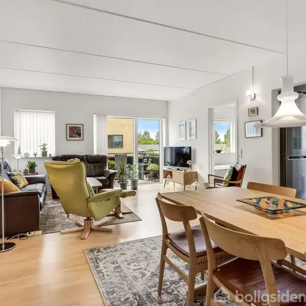 Stue med spisebord og stole, sofaer og en lænestol i lyse farver. Store vinduer giver naturligt lys. Et åbent køkken ses til højre. Udendørs balkon i baggrunden.