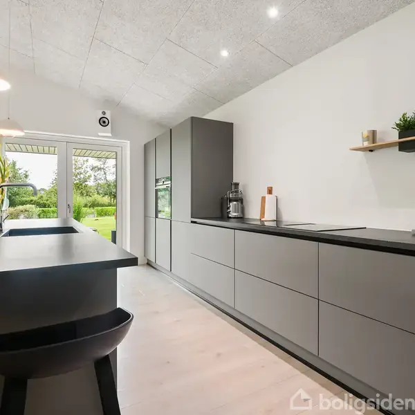 Et moderne køkken med grå skabe og en sort køkkenø. To sorte barstole står ved øen. Glasdøre fører ud til en grøn have. Loftet har indbyggede lys.