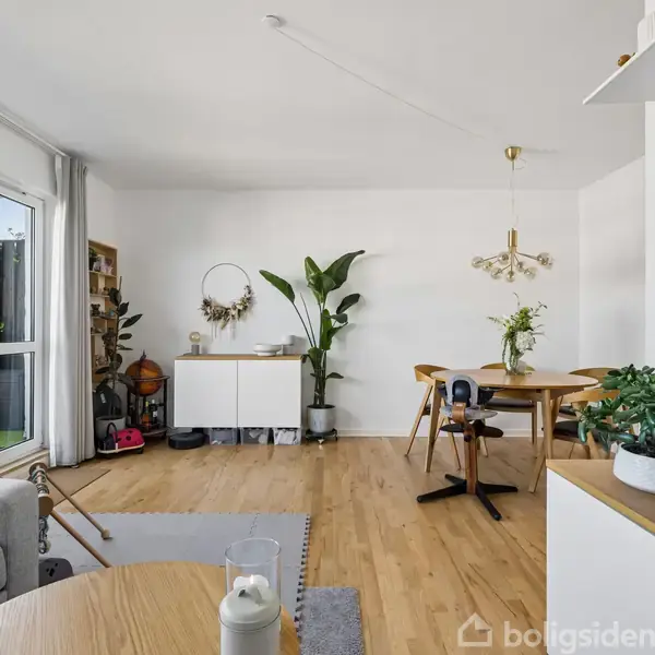 Stue med trægulv, spisebord med stole og grønne planter i hjørnet. Stor skænk med dekorationer. Tv til højre. Stort vindue med udsigt til balkon. Hyggelig, moderne indretning.