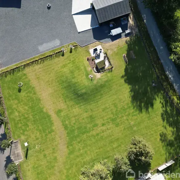 En stor, velholdt græsplæne med en terrasse og belægningssten i et hjørne. Terrassen indeholder møbler, omgivet af en bygning til venstre og træer til højre med en vej.