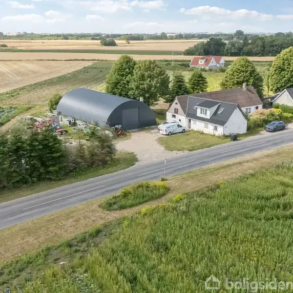 En landlig ejendom med huse og en stor buet lade ligger ved en vej omgivet af marker og træer. Flere biler er parkeret på gårdspladsen.