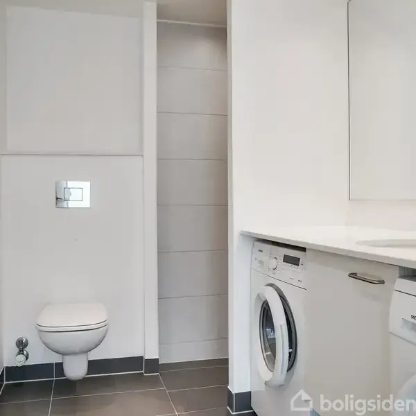 Vaskemaskine og tørretumbler står under en vask i et moderne badeværelse med rummelig bruser. Toilet er monteret på væggen ved siden af en flisebeklædt bruseniche.