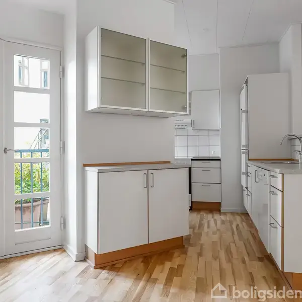 Et lyst køkken med trægulv. Køkkenskabe med glaslåger og hvide skabe står langs væggen. Franske døre åbner mod en balkon med grønne planter udenfor.