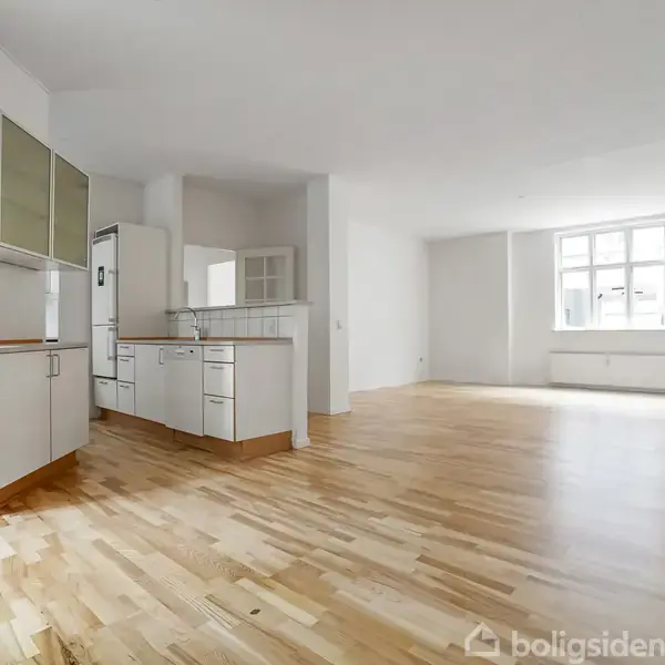 Et rummeligt, lyst køkken-alrum med træparketgulv og hvide vægge. Køkkenet har hvide skabe og indbyggede apparater. Store vinduer i baggrunden giver naturlig belysning til den åbne stue.