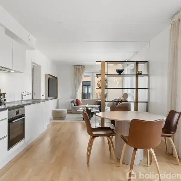 En moderne, lys stue-køkken kombination med hvidt køkken, rund spisebord og fire træstole. Stue med sofa og TV bagved, store vinduer med lyse gardiner.