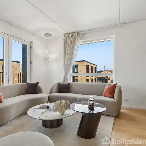 To beige sofaer står i stuen foran store vinduer. Et rundt, moderne sofabord står på et tæppe. Reoler med bøger og dekoration er til venstre i rummet.