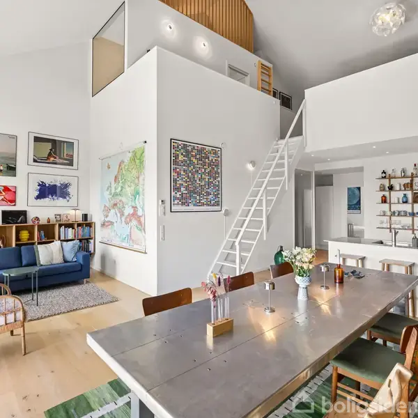 Et moderne, lyst rum med højt til loftet. Et langt spisebord står i forgrunden. En blå sofa og kunst pryder væggen. En trappe fører op til et loftsrum.
