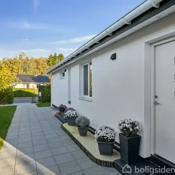 En hvid husfacade med flere dekorerede blomsterkrukker står på en flisebelagt gangsti. I baggrunden ses en velplejet græsplæne og træer under en klar blå himmel.