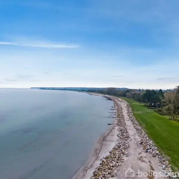 Kystlinje strækker sig langs roligt blåt vand, med en stenstrand til højre og frodig grøn vegetation længere ind mod land. Skyfri blå himmel danner en fredelig baggrund.
