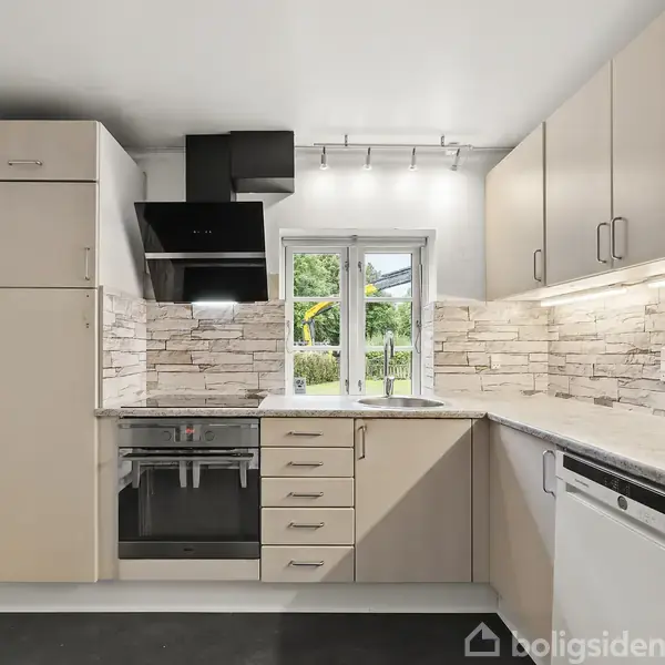 Køkken med beige skabe og stenvæg bagved. Ovnen og komfuret er integreret, og en sort emhætte hænger over. Et vindue giver udsigt til en grøn have.