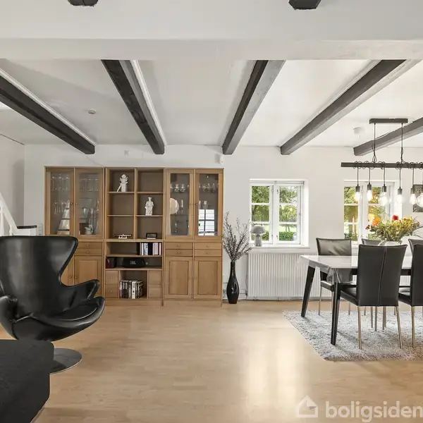 Sort lænestol står stille i en moderne stue med træbjælker i loftet, ved siden af et spisebord og vitrineskab. Store vinduer giver naturligt lys.