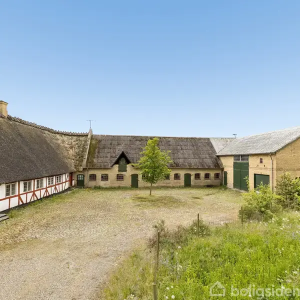 Gårdebygning med stråtag og bindingsværk står stille på en gårdsplads. Enkelt træ i midten, omgivet af græs og sten. Klar blå himmel i baggrunden.
