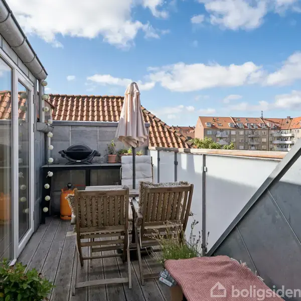 Terrassen har en træstol og bord med en parasol, omkranset af glasvægge. En grill står ved siden af, og bygninger er synlige i baggrunden under en klar blå himmel.