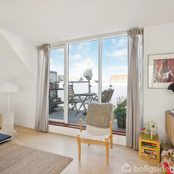 Stue med lys træstol og farverig pude foran glasdør til balkon. Sofa til venstre, træbord foran TV-reol med bøger og lamper til højre. Udsigt til himmel.