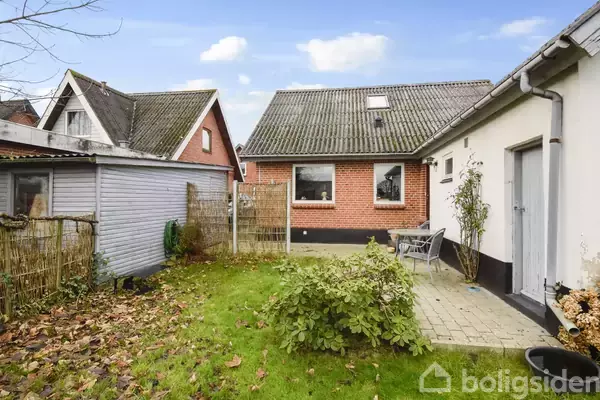 En lille, velholdt baghave med græsplæne og flisebelagt terrasse omgivet af lave hegn. I baggrunden står et rødstenshus med et skråt tag og et mindre skur til venstre.