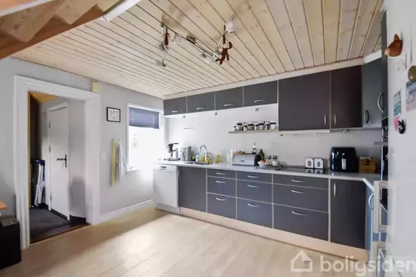 Køkken med grå skabe under og over en hvid bordplade, indeholder køkkenmaskiner og krydderiglas; lyst trægulv og loft. Rummet har en dør til en anden del af huset.