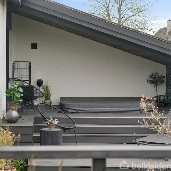 En overdækket terrasse med en hævet platform, indeholder moderne sorte havemøbler og planter. Terrassen støder op til et hvidt hus med trærammer omkring vinduerne.