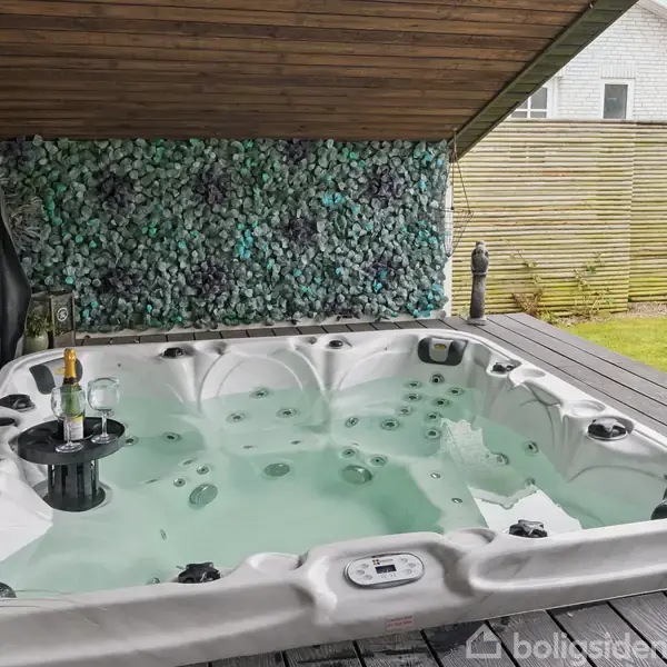 Udendørs boblebad fyldt med vand, placeret på en terrasse, med en flaske champagne og glas på en flydende bakke. I baggrunden ses en hæk og et hus.