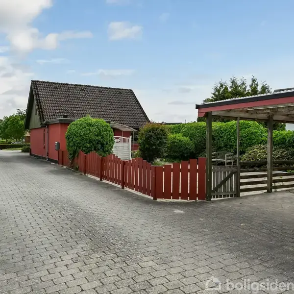 En rød bygning med mørkt tag står ved siden af en flisebelagt vej. Omkringliggende buske og et lille plankeværk skaber en hyggelig atmosfære.