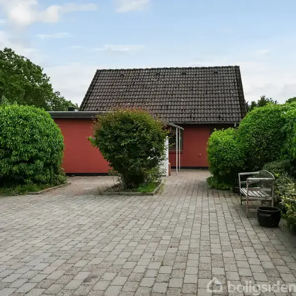 Rødt hus med skråt tegltag står i baggrunden. Foran ses en brolagt indkørsel omkranset af grønne buske og træer. En bænk og planter ses til højre.