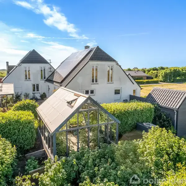 En hvid villa med skråt tag og flere vinduer står ved siden af et drivhus omgivet af tætte, grønne buske; en græsplæne strækker sig ud i det solrige landskab.
