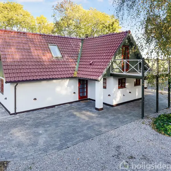 Et charmerende hus med rødt tegltag og hvid facade står omgivet af grønne træer. Huset har en balkon, og en flisebelagt indkørsel leder hen til hoveddøren.