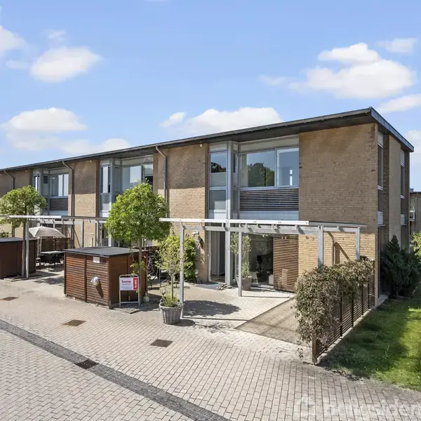 Rækkehus i moderne stil, står roligt med store vinduer og beige murstensfacade. Det er omgivet af en flisebelagt gangsti, små træer og grønne områder under en blå himmel.