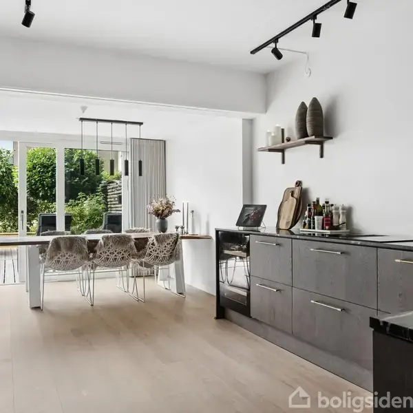 Et moderne køkken med grå skabe; rummet åbner op til en spiseplads med glasbord og fire stole. Store glasdøre i baggrunden fører ud til en frodig have.