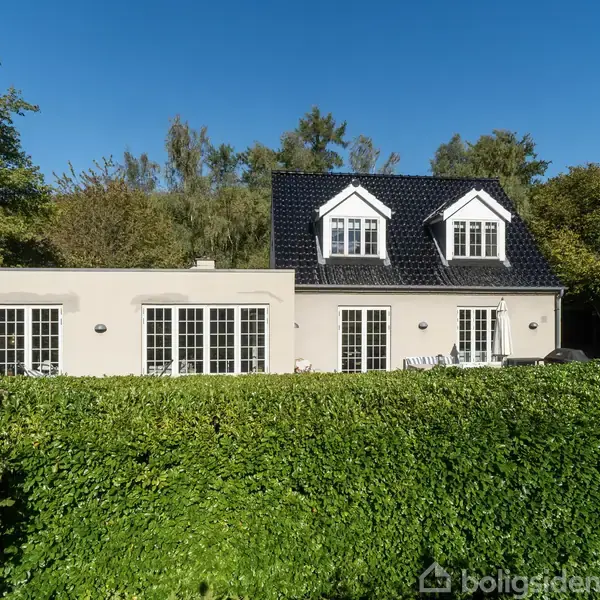 Hus med hvid facade og sort tag står bag en velplejet, høj hæk. Det har store vinduer og en rolig have omgivet af træer.