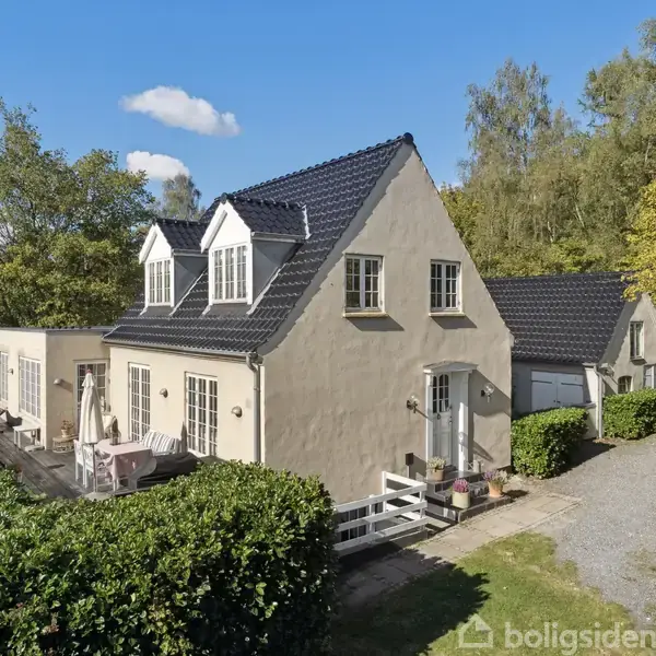 Et hvidt hus med sorte tage står omgivet af grønne buske og træer i en landlig omgivelser. Der er en terrasse med møbler og en grusvej ved siden af.