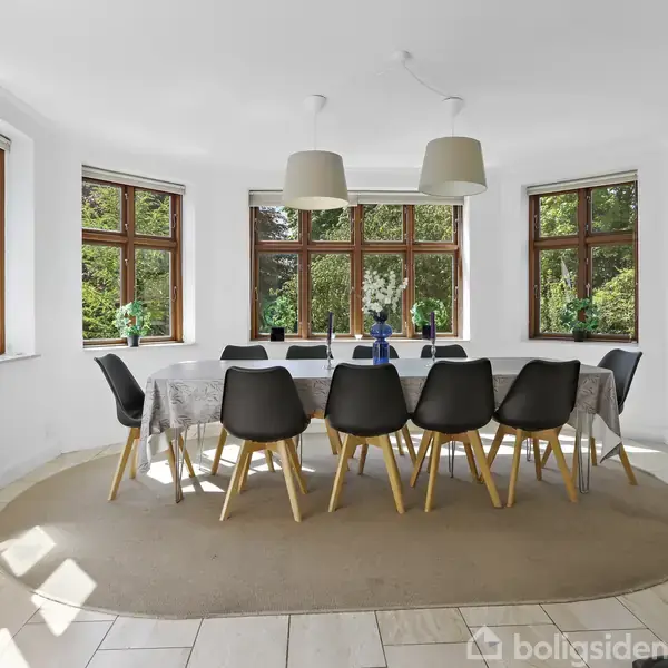 En stor spisestue med et langt bord dækket med hvide duge og omgivet af sorte stole. Rummet er lyst med store vinduer, der giver udsigt til grønne træer udenfor.