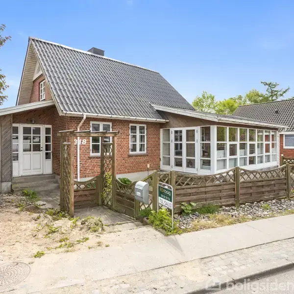 Et rødt murstenshus med hvide vinduer, omkranset af et hegn. Foran huset ses en postkasse og skiltet "Til salg". Huset er beliggende i et boligkvarter.